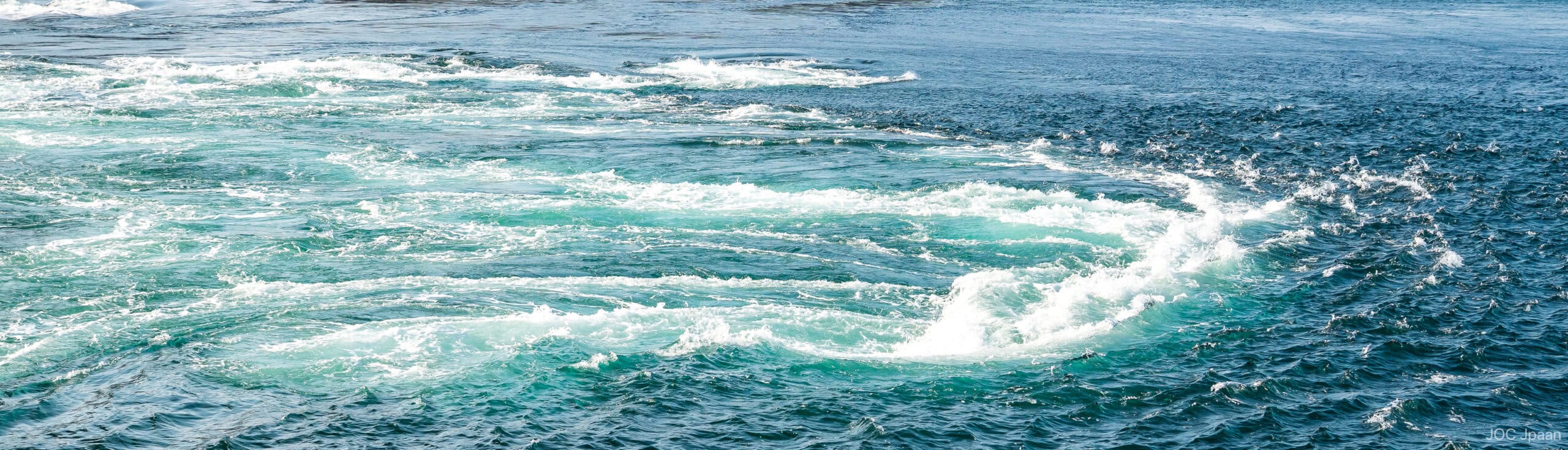 Churning turquoise and deep blue ocean water forms swirling currents and white foamy waves on the surface.